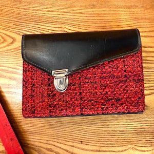 May include: A small, rectangular clutch with a black leather flap and a red woven body. The clutch has a silver clasp and is likely used for carrying small items like a wallet or phone.