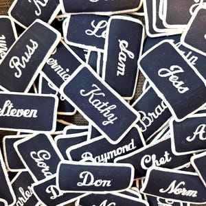 May include: A collection of navy blue name tags with white embroidered names. The tags are rectangular with rounded corners and a white border. Names include Kathy, Steven, and Jess.