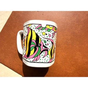 May include: White ceramic mug featuring a colorful, whimsical design of two tropical fish. The fish have yellow, black, and pink stripes, with green and pink accents. The mug has a white handle and is set against a brown surface.