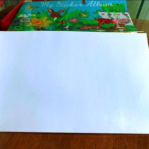 May include: An open sticker album with blank white pages. The cover of the album is visible, featuring a colorful illustration of a garden scene with a rabbit, flowers, and butterflies. The album is on a wooden surface.