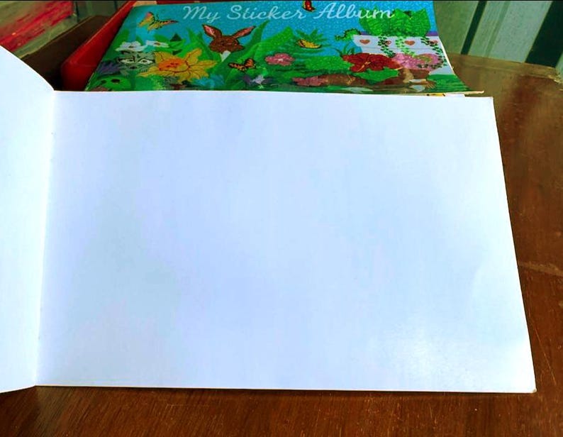 May include: An open sticker album with blank white pages. The cover of the album is visible, featuring a colorful illustration of a garden scene with a rabbit, flowers, and butterflies. The album is on a wooden surface.