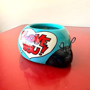 May include: A whimsical, ceramic ladybug-shaped planter. The turquoise body features a white heart with the words "I LOVE YOU!" in red. Black antennae and a black head complete the design. The planter sits on a red surface.