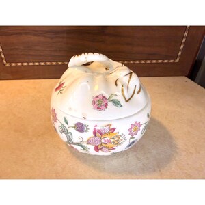 May include: A round white ceramic sugar bowl with a lid featuring a floral pattern in shades of pink, yellow, green, and purple. The lid has a sculpted handle and gold-colored accents. The body of the bowl is decorated with various botanical illustrations.