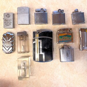May include: A collection of twelve assorted metal and plastic lighters in various shapes and finishes. One silver lighter features text including 'Joe Zielinski Rev. Co.' and 'Leavenworth, Kansas'. Others include a shell-patterned silver lighter, a black and gold rectangular lighter, and a rectangular lighter with a landscape graphic.