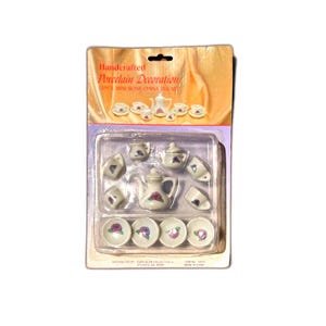 May include: A miniature porcelain tea set, featuring a teapot, cups, saucers, and sugar bowls. The set is white with a floral design and is packaged in a plastic container. The packaging reads "Handcrafted Porcelain Decoration".