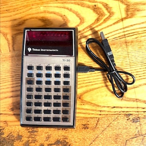 May include: A vintage TI-30 calculator with a black and silver frame, featuring numerous buttons and a red display screen. The calculator is connected to a black USB cable. The Texas Instruments logo is visible.