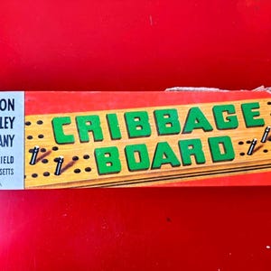 May include: Vintage cribbage board game by Milton Bradley Company. The box is red and features the words "CRIBBAGE BOARD" in large green letters. The board is made of wood and has holes for pegs. The box also includes the company's name and location.