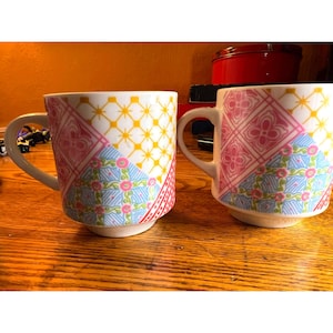May include: Two white ceramic mugs with colorful patchwork designs. Each mug features sections of pink, blue, and yellow patterns. The mugs have curved handles and sit on a wooden surface.