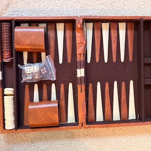May include: A brown leather-like backgammon set in an open case with a dark brown felt interior. The board features alternating brown and white triangular points. Included are two brown dice cups, a stack of white checkers, a stack of brown checkers, and a plastic bag containing four white dice and one doubling cube with the number 64.