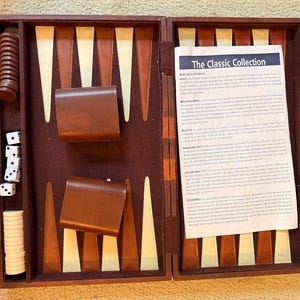 May include: A wooden backgammon board game set featuring a brown and cream triangular point pattern, two brown leather dice cups, white and brown checkers, four white dice, and an instruction sheet titled The Classic Collection Backgammon.