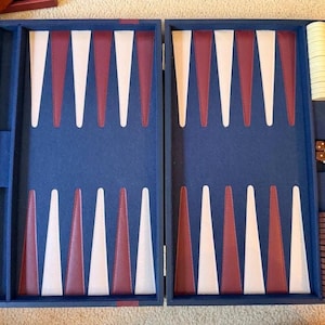 May include: A blue backgammon board with alternating white and maroon triangular points. The set includes a stack of white checkers, a stack of brown checkers, and two white dice with brown pips, all arranged within the storage compartments.