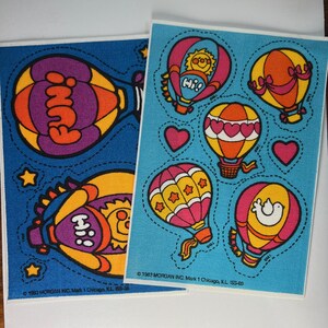 May include: Two sheets of colorful stickers featuring hot air balloons with various designs. The stickers are on a blue background and have a white border. The text on the stickers reads "Fun!" and "Hi!"
