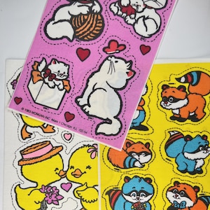 May include: Three sheets of stickers featuring various animals. The first sheet is pink and features white cats with yarn balls, hearts, and a cat carrying a kitten. The second sheet is white and features a yellow duck couple holding flowers and hearts. The third sheet is yellow and features raccoons with flowers and a bow tie.