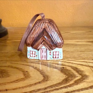 May include: A ceramic cottage ornament with a brown thatched roof and a light blue base. The cottage has a pink door and windows with floral accents. A brown ribbon is attached for hanging.