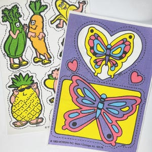 May include: A set of vintage peel and stick stickers featuring a pineapple, a banana, a carrot, celery, and two butterflies. The stickers are on a white and purple background. The stickers are from 1983 Morgan Inc. Mark I Chicago, Ill. 553-70.