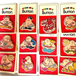 May include: A collection of vintage "Human Bean" buttons on red and white cards. Each button features a cartoon bean character with various sayings, including "Baby me" and "This Human Bean needs love!"