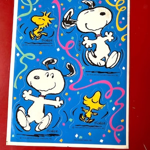 May include: A sheet of Snoopy and Woodstock stickers on a blue background with colorful confetti and streamers. The stickers feature Snoopy in various poses and Woodstock. The text "SCHULZ" and copyright information are visible.