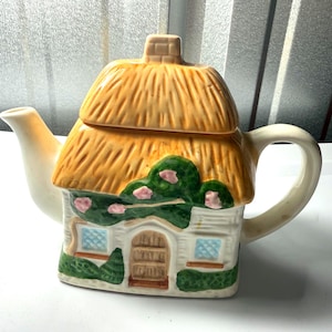 May include: A ceramic teapot shaped like a cottage. The teapot features a thatched roof, a chimney, and a house facade with windows and a door. Green bushes and pink flowers decorate the house. The teapot is cream-colored with yellow and green accents.