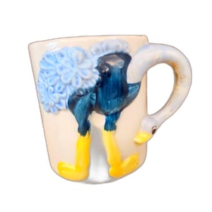 May include: A whimsical ceramic mug featuring a colorful ostrich design. The mug is primarily cream-colored, with the ostrich depicted in shades of blue, yellow, and white. The handle is shaped like a swan's neck and head, adding to the unique design.