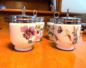 May include: Four white ceramic egg coddlers with shiny chrome lids and metal ring handles. The jars feature colorful botanical and animal illustrations, including a large purple flower, a rabbit holding a carrot, a butterfly, and a mouse.