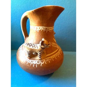 May include: A brown clay pitcher with white painted designs and three small animal figures. The pitcher has a wide mouth and a handle.