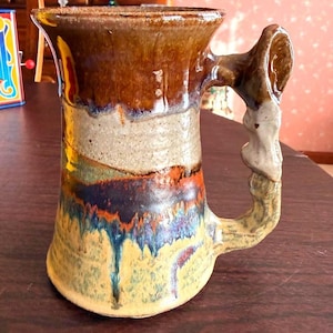 May include: Handmade ceramic mug with a unique, textured design. The mug features a brown, tan, and blue color scheme with a distinctive handle. The mug's exterior has a glossy finish and a flared rim, perfect for enjoying hot beverages.