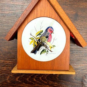 May include: A wooden birdhouse-shaped clock with a round face featuring a colorful bird illustration. The clock has a natural wood finish and a small base. The clock hands are gold-colored. The text "CHAFFINCH" is printed on the clock face.