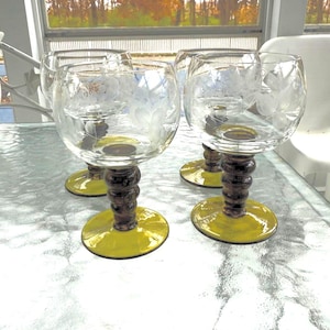 May include: Four clear wine glasses with etched floral designs. The stems are dark brown with a bulbous shape, and the bases are a bright yellow. The glasses are arranged on a reflective surface.