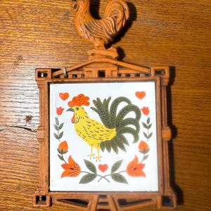 May include: A vintage trivet featuring a rooster design. The trivet has a square tile with a yellow rooster, red comb, and green tail, surrounded by orange flowers and hearts. The frame and rooster figurine are a rusty orange color.