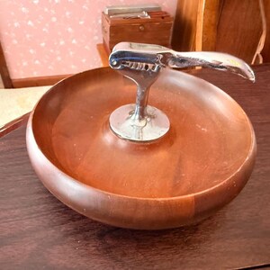 May include: A wooden nut bowl with a built-in chrome nutcracker. The bowl is round and has a rich, reddish-brown color. The nutcracker is attached to the center of the bowl and is made of shiny chrome.