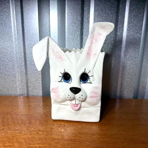 May include: A white ceramic bunny-shaped container with blue eyes, pink cheeks, and a black nose. The bunny has long ears with pink accents and a smiling mouth. The container is designed to resemble a paper bag.