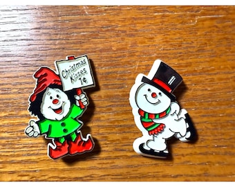 May include: Two Christmas-themed decorative pins. One is a smiling elf holding a sign that reads "Christmas Kisses 1¢". The other is a snowman wearing a top hat and scarf.