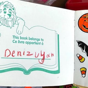 May include: Open book with a lion illustration and the text "This book belongs to Ce livre appartient &agrave; Deniz Uyan". The right page has Halloween-themed stickers, including a jack-o'-lantern, bat, and candy corn.