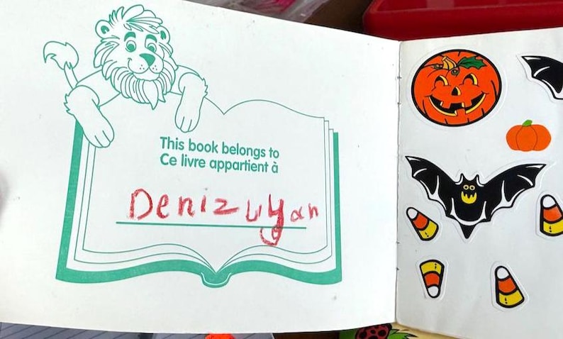 May include: Open book with a lion illustration and the text "This book belongs to Ce livre appartient &agrave; Deniz Uyan". The right page has Halloween-themed stickers, including a jack-o'-lantern, bat, and candy corn.