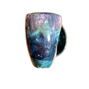 May include: A ceramic mug featuring a glossy glaze with a blend of deep navy blue, teal, and turquoise tones. The upper rim displays a textured, crystalline pattern in shades of light purple and white that drips down into the darker blue body.