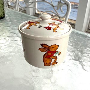 May include: A cream-colored ceramic jar with a lid, featuring illustrations of brown rabbits wearing red scarves. The jar has a small knob on top and holes in the lid. The rabbits are holding gifts, creating a festive theme.