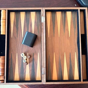 May include: A wooden backgammon board game set featuring a folding case with a brown and tan triangular point pattern. The set includes two stacks of light and dark wooden checkers, a black dice cup, and four small white and brown six-sided dice.