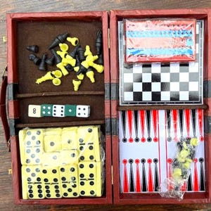 May include: A red, hinged travel game set with multiple games. The set includes chess pieces, dice, dominoes, and a backgammon board. The chess board is black and white. The dominoes are yellow with black dots. The backgammon board has red and black triangles.
