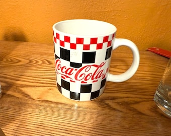 May include: White ceramic Coca-Cola mug with a red, black, and white checkered pattern. The Coca-Cola logo is in red script. The mug has a white handle and is on a wooden surface.