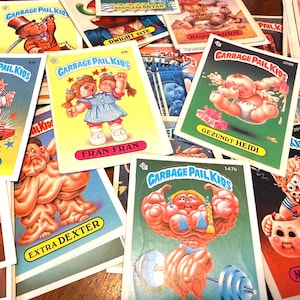 May include: A collection of Garbage Pail Kids trading cards. The cards feature cartoon characters with exaggerated features and humorous names. The cards are colorful, with various designs and text, including names like "Fran Fran" and "Extra Dexter."