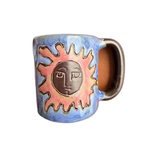 May include: A ceramic mug with a blue speckled glaze featuring a central dark brown sun motif with a human face and pink rays. The mug has a sturdy handle and a smooth, rounded cylindrical body, showcasing a handcrafted aesthetic with visible texture.