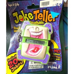 May include: A green and purple plastic toy keychain shaped like a mouth with a red tongue sticking out. The keychain has a button that says "Try Me!" and a screen that says "Why did the..." The keychain is packaged on a card with the words "Joke Teller" and "20 Jokes!" printed on it.