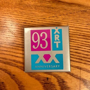 May include: A square, silver-colored pin with a pink and blue design. The pin features the number "93" in pink, the letters "XRT" in blue, and the word "ANNIVERSARY" in blue. A pink diamond shape is also present.