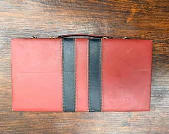 May include: A rectangular, red case with a black stripe down the center and two black stripes on either side. The case has a handle and gold-colored hardware. The case is sitting on a wooden surface.