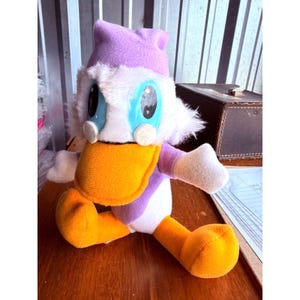 May include: A plush doll of a cartoon duck character, featuring a large orange beak, blue eyes, and a purple hat. The doll is primarily white, purple, and orange, with white fluffy accents around the face. The doll is sitting on a wooden surface.