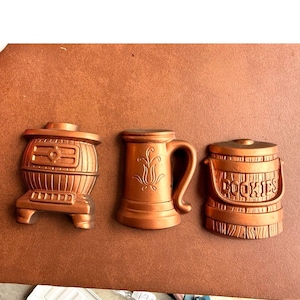 May include: Three copper-colored decorative wall hangings: a stove, a beer mug with a floral design, and a cookie jar with the word "COOKIES". The items are arranged on a brown surface.