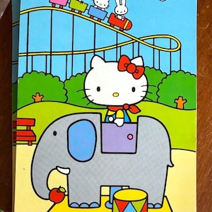 May include: A Hello Kitty-themed children's activity book. The cover features Hello Kitty riding an elephant, with the text "1 Play Elephant 50¢". The background includes a roller coaster with other cartoon characters. The book measures 240 mm x 130 mm.