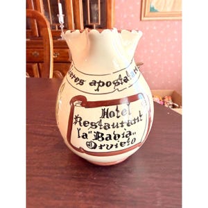 May include: Cream-colored ceramic pitcher with a ruffled rim. The pitcher features brown lettering and a banner that reads "Hotel Restaurant La Bahia Orvieto". Decorative item.