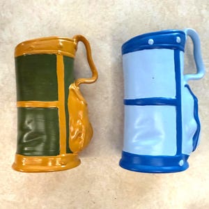 May include: Two ceramic mugs with handles. One is green and gold, the other is light blue and dark blue. The mugs have a unique, barrel-like shape with a curved handle. The mugs are on a light-colored surface.