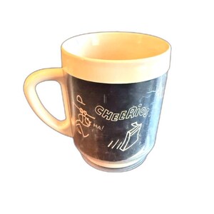 May include: A vintage Cheerios mug with a cream-colored rim and handle. The mug's body is black with white text and illustrations, including the word "Cheerios" and a box of cereal. The mug has a classic, rounded shape.